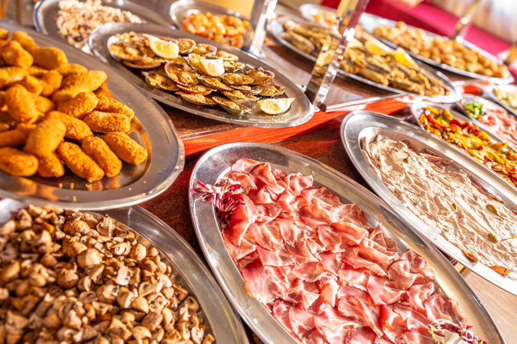 Un buffet di antipasti assortiti su vassoi d'acciaio, tra cui salumi, funghi e crostini.