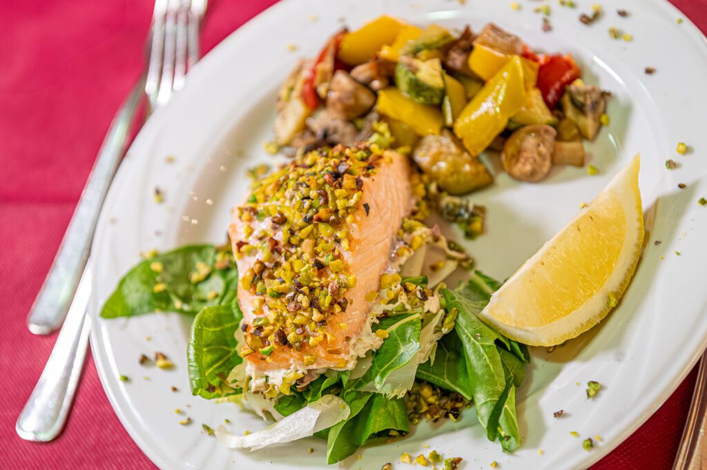 Filetto di salmone in crosta di pistacchi con contorno di verdure e fetta di limone su un piatto bianco.