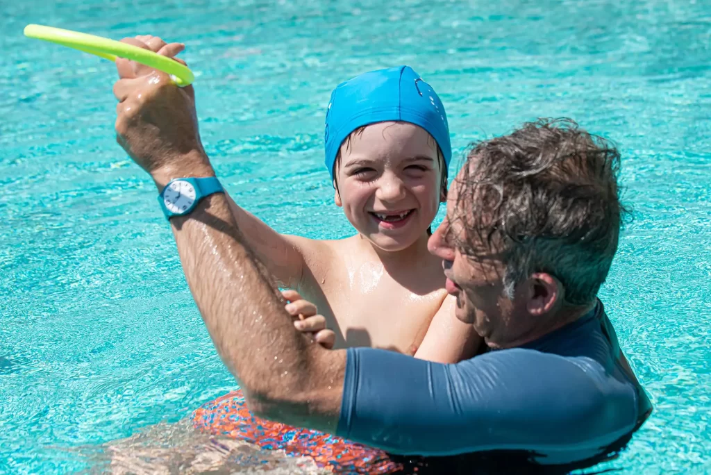 Un uomo gioca felicemente in piscina con un bambino che indossa una cuffia blu.