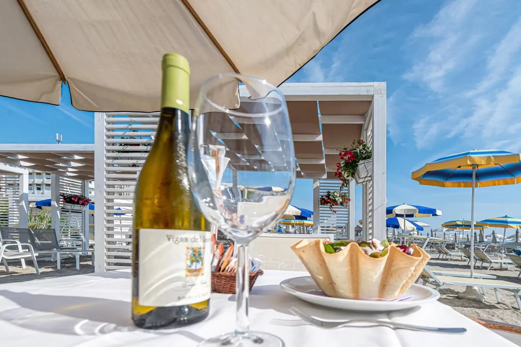 Bottiglia di vino e bicchiere su tavolo con ombrelloni e lettini al mare.