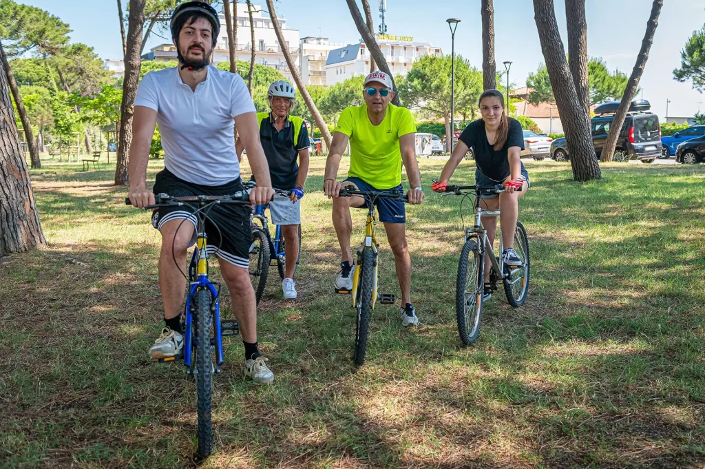 Quattro persone in tenuta sportiva pedalano in un parco alberato su biciclette.