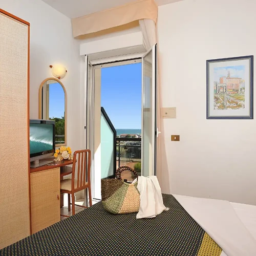 Camera d'albergo luminosa con balcone vista mare, arredamento semplice e televisore.