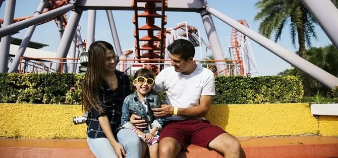 Famiglia sorridente seduta davanti a una montagna russa in un parco divertimenti.